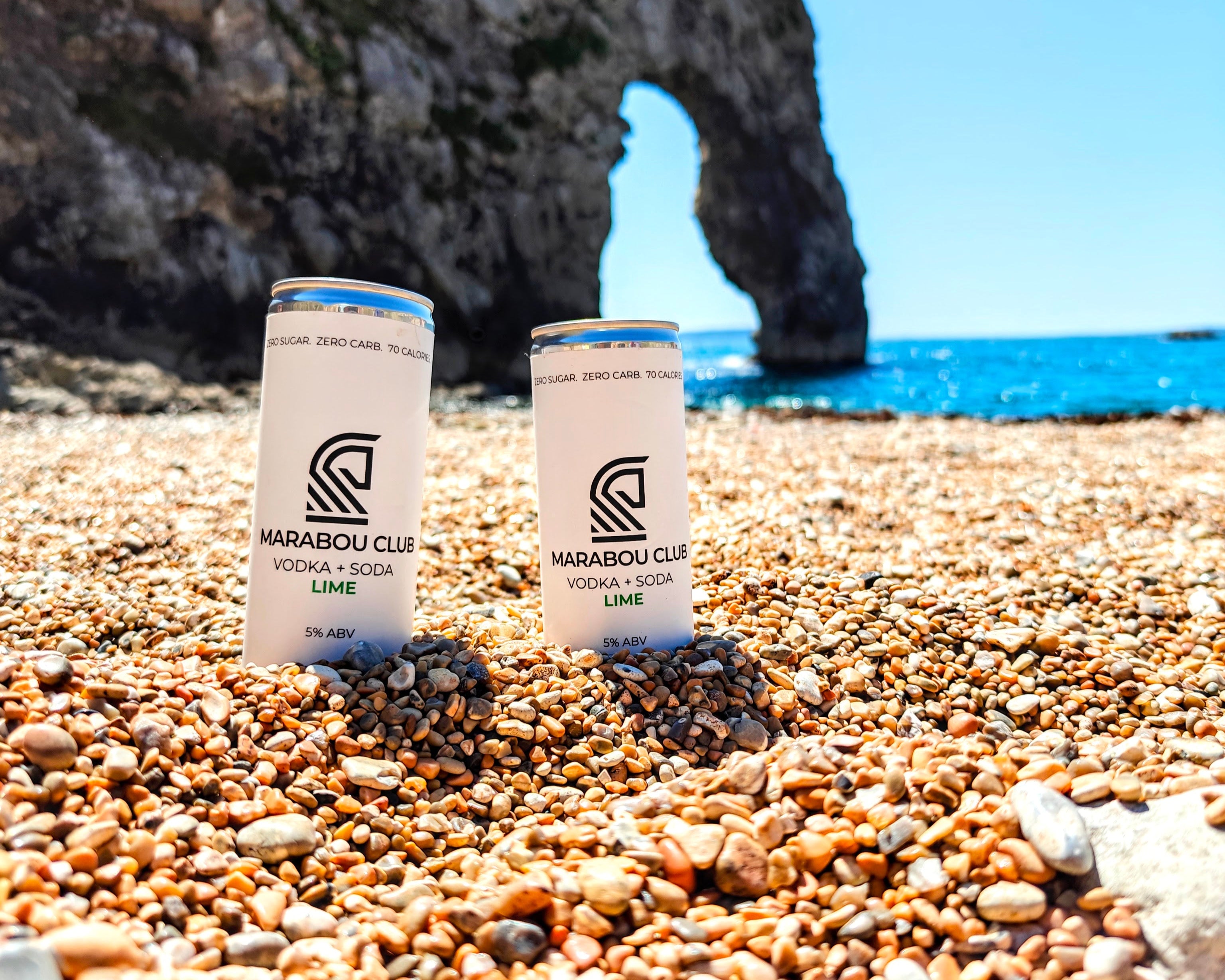 Two Marabou Club cans on a pebbly beach with a rock formation and blue sky in the background.