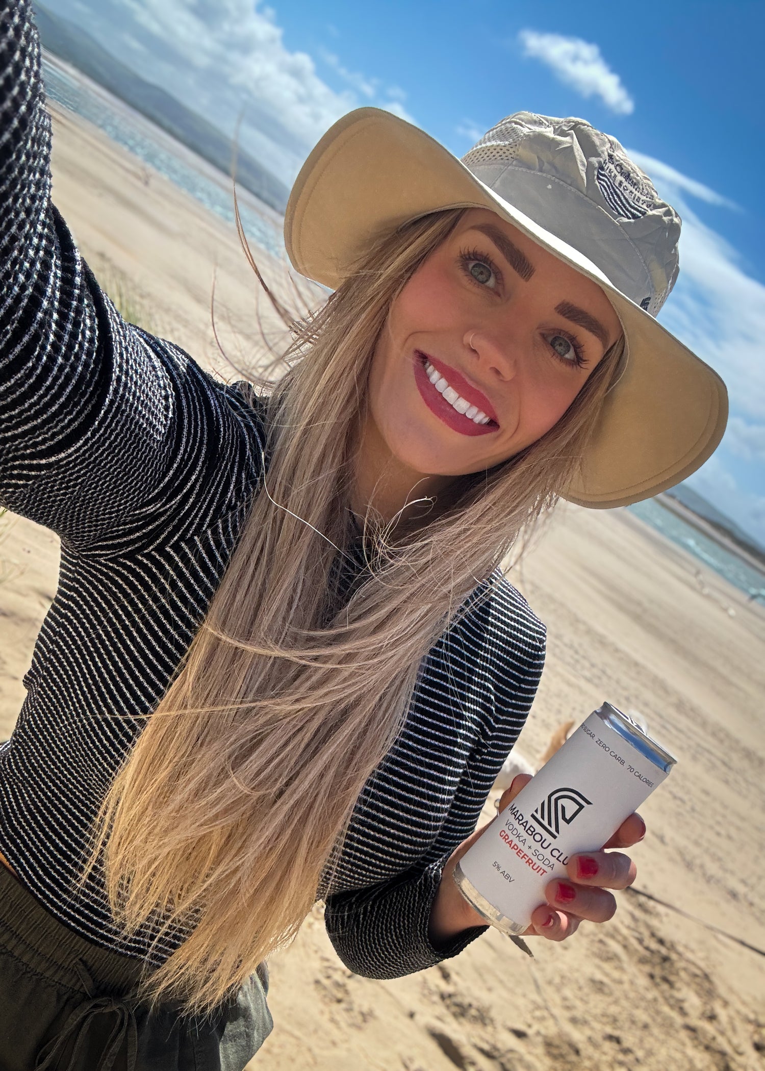 Woman on a beach holding a product, with a clear sky and ocean in the background